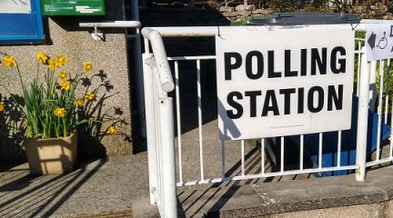 A sign saying 'polling station' on railings leading to the door of a village hall, with a tub of daffodils to the left