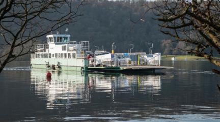 Windermere ferry sailing on calm lake with tree branches to left and right.