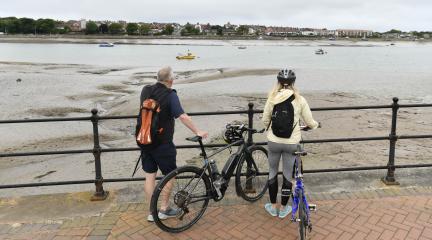 A man and a woman standing with bikes at railings looking over Walney Channel towards Walney