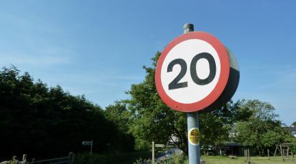 A 20mph sigh at the side of a road with a playground in the background and a hedge to the side