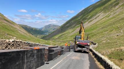 A yellow digger in a valley road with hills to either side leading to blue sky.