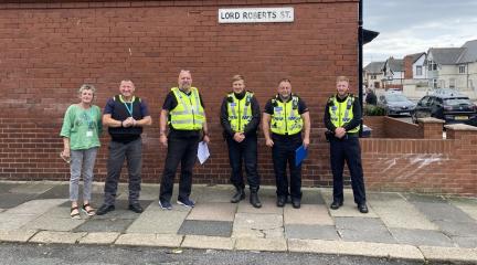 Six people from the police and the council in high visibility clothing, standing in front of a house wall.