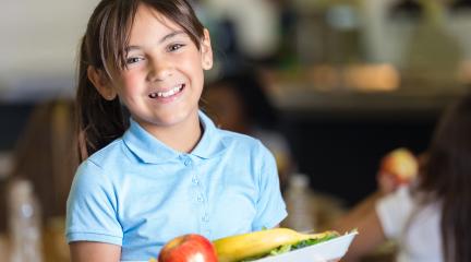 A girl in school uniform holding a school dinner tray, with an apple and banana on top.