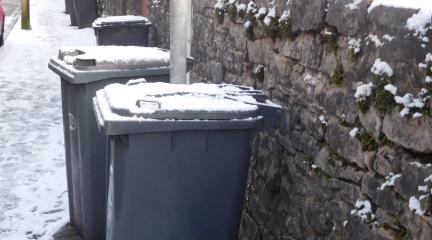 A line of grey waste bins on a pavement covered in snow.