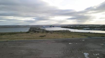 A flat expanse of land with scrubland and docks beyond.