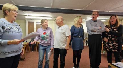 Six actors standing in a line holding scripts rehearsing a pantomime.