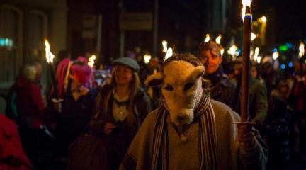 A line of people carrying lit torches at night as part of a parade, one of them wearing an animal mask.