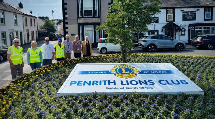 A group of people standing beside a display of yellow and blue flowers with a Penrith Lions Club banner at the centre.