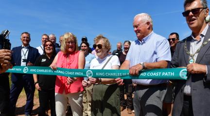 A line of people holding a green ribbon, with a woman in the middle cutting it.