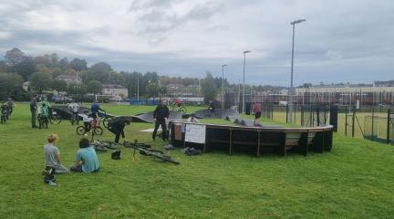 A busy pump track with groups of young people sitting on the grass and people using it.