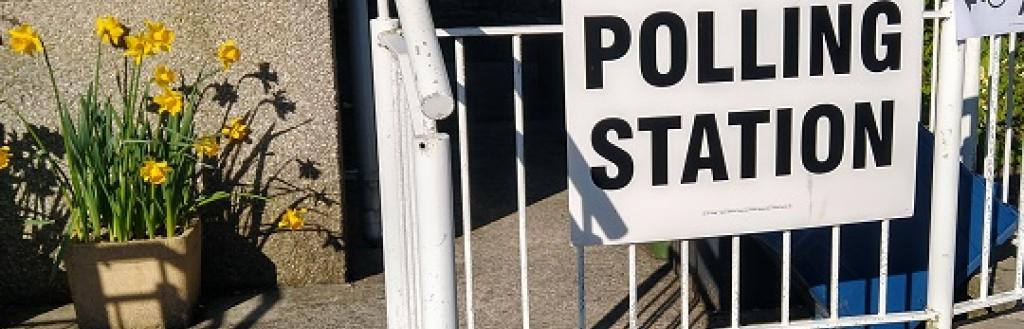 A sign saying 'polling station' on railings leading to the door of a village hall, with a tub of daffodils to the left