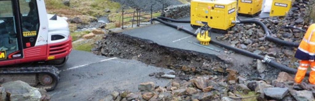 A section of road at Kirkstone Pass with a trench dug out of it and a digger.