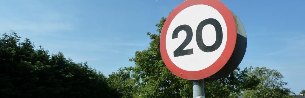 A 20mph sigh at the side of a road with a playground in the background and a hedge to the side