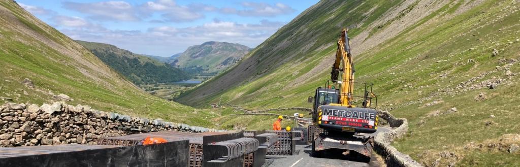 A yellow digger in a valley road with hills to either side leading to blue sky.