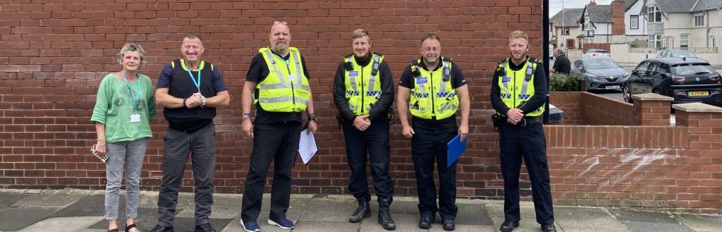 Six people from the police and the council in high visibility clothing, standing in front of a house wall.