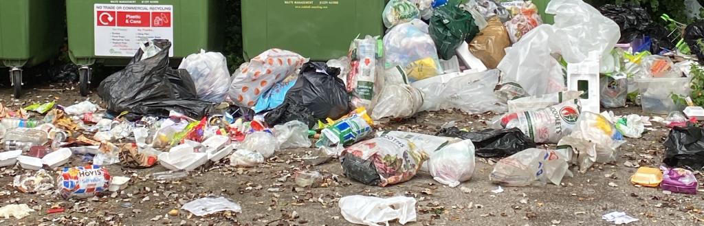 Rubbish piled up and strewn in front of green recycling banks.