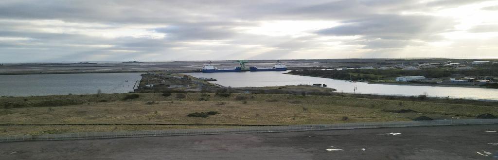 A flat expanse of land with scrubland and docks beyond.