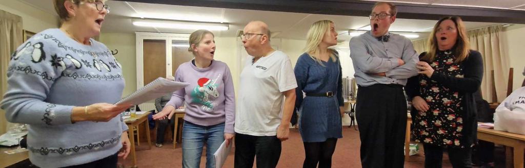 Six actors standing in a line holding scripts rehearsing a pantomime.