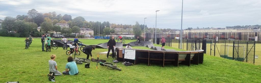 A busy pump track with groups of young people sitting on the grass and people using it.jpg