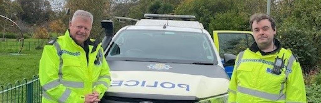 Two CCTV operators in hi-vis yellow jackets standing beside a police car in a park. CCTV camera in the background.