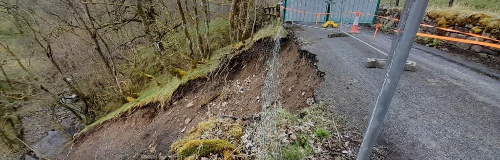 Picture showing landslip at Cowgill, with fencing around washed away section of road and steep drop to a stream below