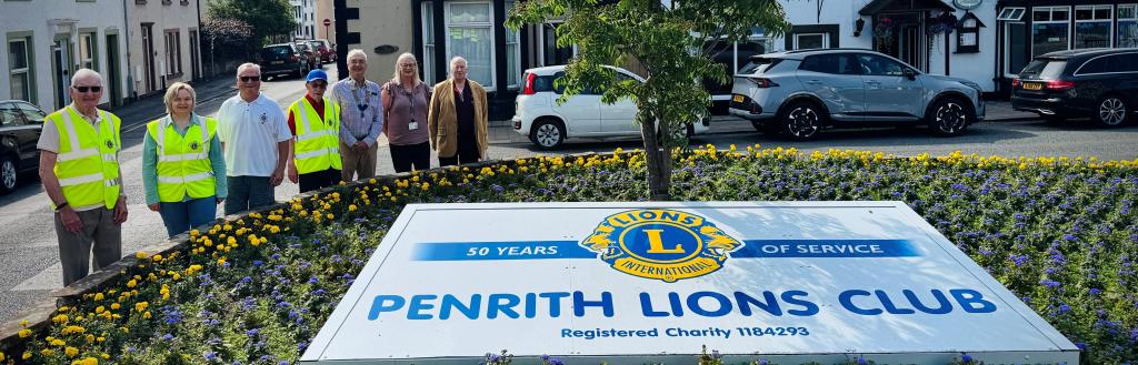 A group of people standing beside a display of yellow and blue flowers with a Penrith Lions Club banner at the centre.