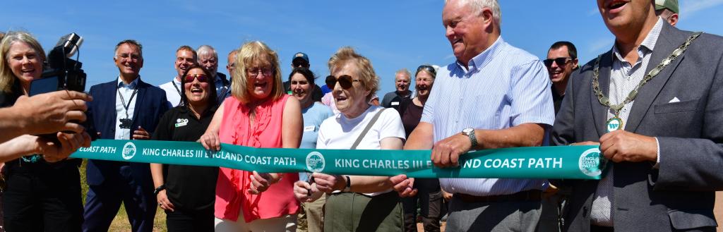 A line of people holding a green ribbon, with a woman in the middle cutting it.