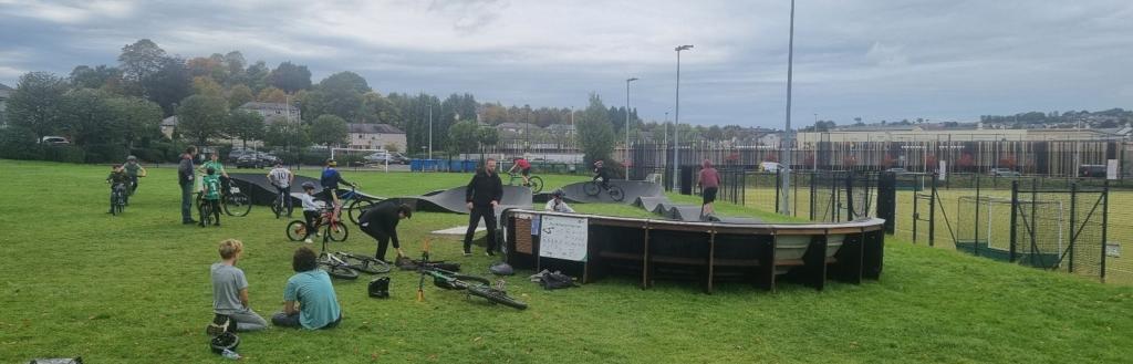 A busy pump track with groups of young people sitting on the grass and people using it.